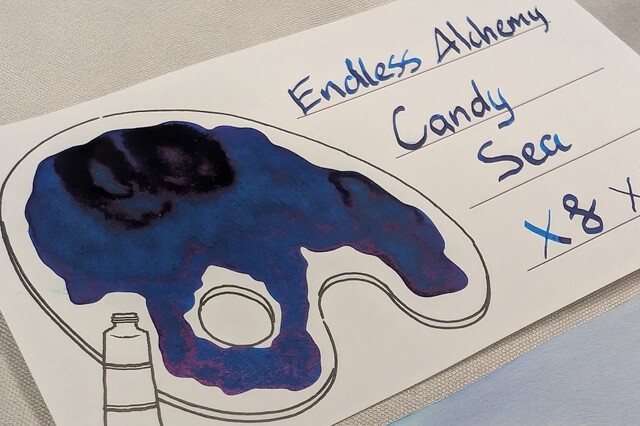 Even lower angle photo an ink swatch card.  Overall the ink is a deep, dark blue with some areas of even darker blue, almost black.  The card is an Iroful swatch card with an outline of a paint palette. Inside the palette is mostly filled in with swirls of midnight blue ink. Some areas with more ink are even darker blue, and areas with even more ink are almost black.