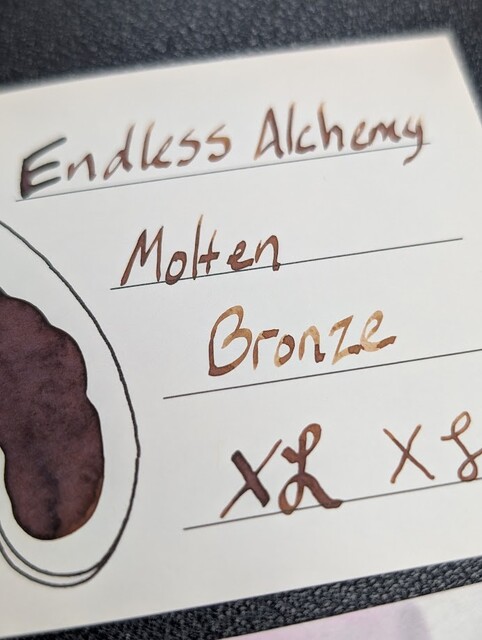 Low angle photo of an ink swatch card with text reading "Endless Alchemy Molten Bronze" followed by an X and curlicue in a stub and EF nib width.  Overall the ink is a medium brown with some shading to a darker hue in parts of the text where more ink settled.