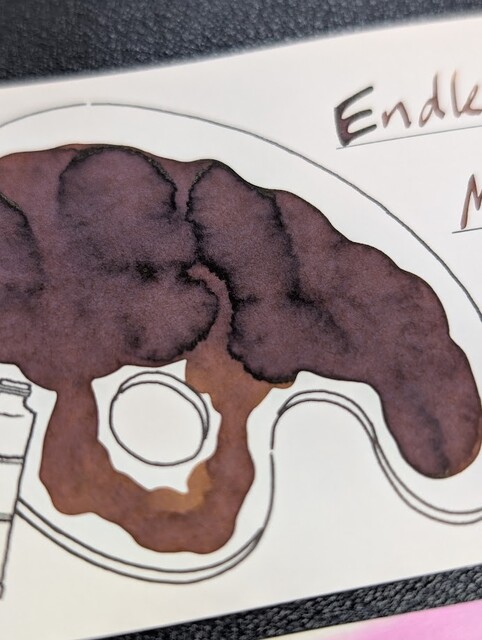 Low angle photo an ink swatch card.  Overall the ink is a medium brown with some shading to a darker hue.  The card is an Iroful swatch card with an outline of a paint palette. Inside the palette is mostly filled in with swirls of brown ink. Areas with less ink are lighter brown, areas with more ink have a little gray tone to them.