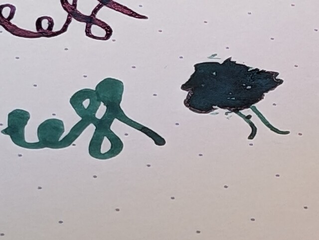 Writing sample on dot grid paper  First line has some swirls. The ink is blue and there is a noticeable reddish sheen on portions of the lines in the writing.  Second line has and some swirls and a tree with the "leaves" filled in with ink. There is a slight yellowish sheen around the edges.