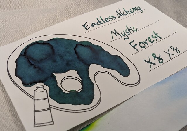 Even lower angle photo an ink swatch card.  Overall the ink is a deep, dark green with very a few darker green areas were more ink has settled. No sheen is visible.  The card is an Iroful swatch card with an outline of a paint palette. Inside the palette is mostly filled in with swirls of hunter green ink. Some areas with more ink are even darker green.
