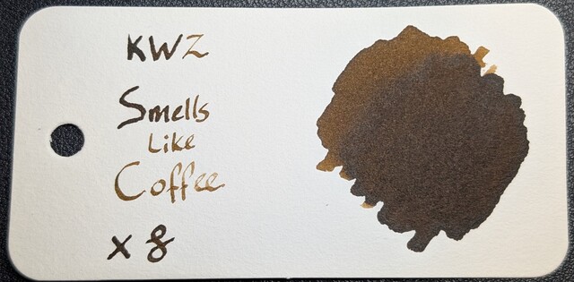 Photo of a col-o-ring swatch card. The ink on the card is a dark brown with some shading so some areas are a little lighter brown. On the right is a large ink blob that is lighter in the upper left and darker in the lower right. On the left is text reading "KWZ Smells Like Coffee" with an X and a curlicue underneath.