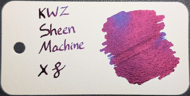 Photo of a col-o-ring swatch card. The ink on the card is a blue almost entirely appearing to be pinkish due to reflective sheen. On the right is a blob of ink with a few places around the edges that have blue showing through. On the right is text reading "KWZ Sheen Machine" with an X and a curlicue underneath.