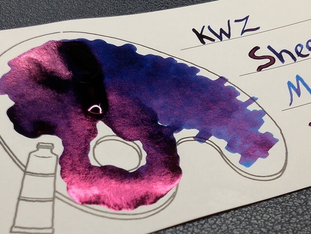 Photo of an Iroful swatch card at an angle with a line drawing of a paint pallette. The ink on the card is a blue with areas in the lower left appearing as pinkish reflective sheen. On the left, inside the line drawing is a blob of ink with a few places around the edges that have blue showing through. On the right is text reading "KWZ Sheen Machine" with an X and a curlicue underneath.