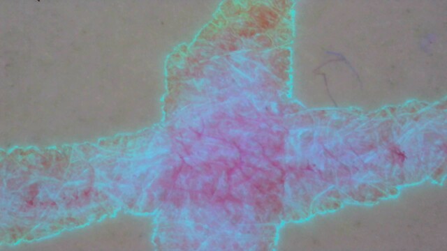 Same as the previous photo, but under a mix of UV and visible light. The center is glowing purple darker fibers glowing more magenta. Some areas of the line are glowing blue with bright blue fibers. Near the edges is it glowing orange and at the very edge is an outline of bright green around everything.