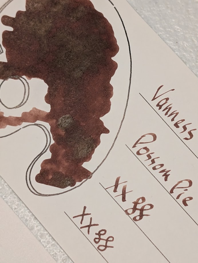 Photo of an Iroful swatch card taken at an angle. It has a writing sample and outline of a palette filled with ink. The ink inside the palette is dark brown with some lighter and darker areas. The darker areas are slightly reflective. The writing sample reads "Vanness Possom Pie" written with a stub nib followed by "XX" and two curlicues written in a stub nib and an F nib. The ink in the writing sample is more of a medium brown with some areas shaded darker.